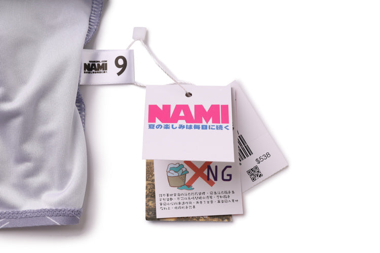 中灰色-MIX&MATCH泳衣上身 <23551488>的產品標籤特寫。主標籤以粗體粉紅字顯示 NAMI,下方則有日文文字,其他標籤則顯示符號、QR 代碼和產品詳細資訊。.
