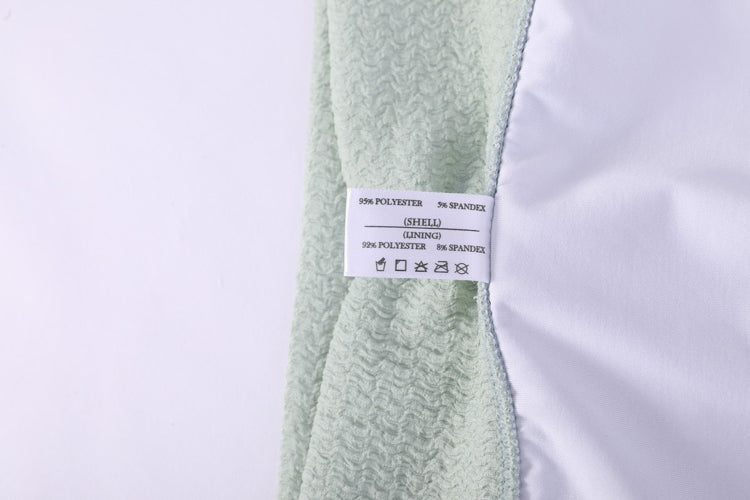 縫在青綠色搭帶MIX&MATCH泳衣下身<24151606>和白色布料上的白色護理標籤特寫,顯示這些修飾身材泳褲的材質細節和洗衣標誌。.