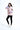 一位女性站在白色背景下，身穿粉紅色撥水防風外套 <20221670-20>、黑色綁腿褲、白色運動鞋。她微笑著用一隻手摸著自己的頭髮。.