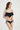 一位身穿 NAMI Bikini 的女性，搭配黑色荷葉邊上衣和 黑色-MIX&MATCH泳衣下身 <24051530>，在素淨的淺色背景下擺姿勢，微微左顧右盼、笑容柔和。.