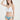 一位女士身穿 NAMI Bikini 的荷葉邊比基尼上衣和天藍色高叉三角MIX&MATCH泳衣下身 <24051623>，在白色背景下擺出造型，她的手舉到頭頂，微微笑著，展示出時尚的高叉比基尼風格。.
