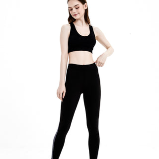 一位身穿黑色緊身長褲抓毛保暖泳衣<26401144-NR01>和白色運動鞋的女性站在純白背景下，低頭微笑，一隻手放在腰間。.