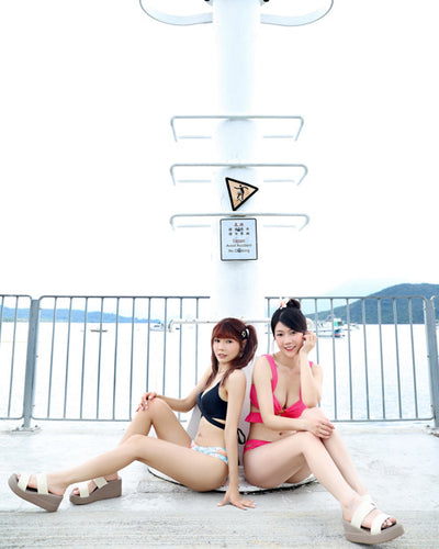 兩位穿著泳衣和平台涼鞋的女士坐在水邊金屬欄杆附近的水泥地上，身後有一根白色柱子和警告標誌。背景是水和遠方的山丘。.