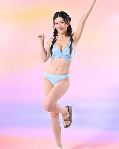 一位身穿淺藍色比基尼和涼鞋的女性單腳站立，舉起一隻手臂微笑。她梳著辮子，在粉紅色和紫色的漸變背景下擺姿勢。.