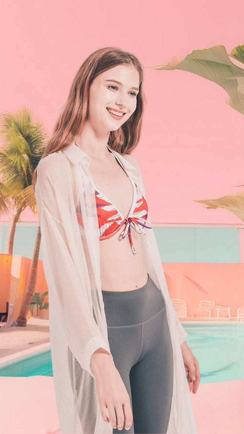 一位身穿白色薄紗罩衫、圖案比基尼上衣和灰色緊身褲的年輕女子站在泳池旁，背景是粉色的建築物、棕櫚樹和粉色的天空。她面帶微笑，目視前方。.
