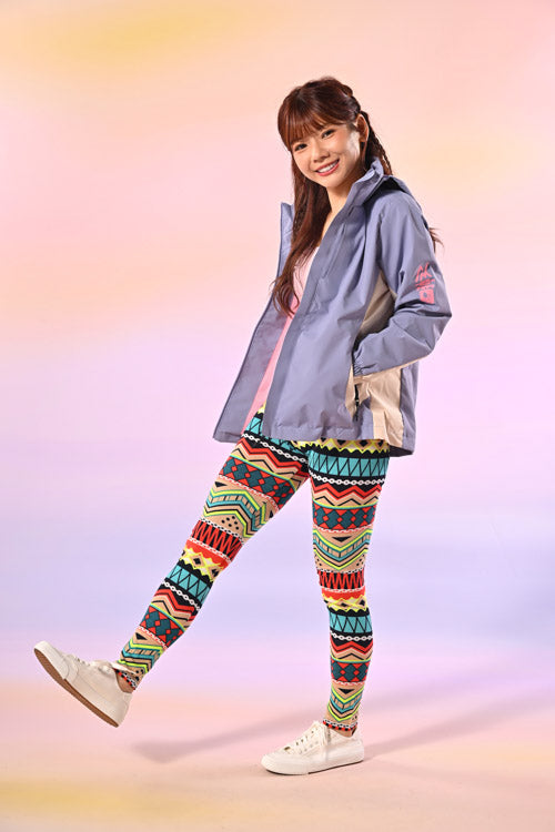 一位面帶微笑的女士單腳站立在粉色漸變背景中，身穿淺紫色夾克、帶有幾何圖案的彩色花紋緊身褲和白色運動鞋。.