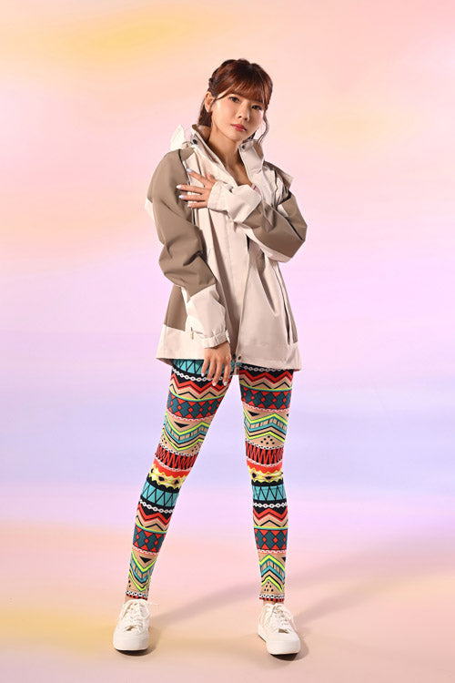 一位女士自信地站在粉色背景下，身穿米色超大外套、帶有幾何圖案的彩色花紋緊身褲和白色運動鞋。她的頭髮梳成馬尾造型。.