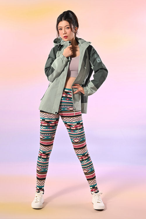 一位女士在粉色漸變背景下自信地擺姿勢，身穿灰色夾克、淺紫色運動胸罩、彩色圖案緊身褲和白色運動鞋。.