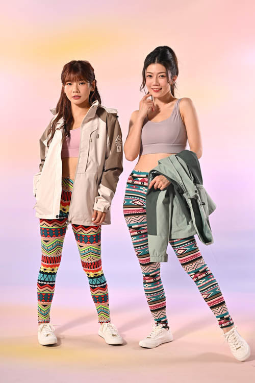 兩個女人並排站著，穿著色彩繽紛的圖案緊身褲、淺色作物上衣和運動鞋。其中一位穿著米色夾克，另一位則拿著綠色夾克。背景是柔和的漸變色。.