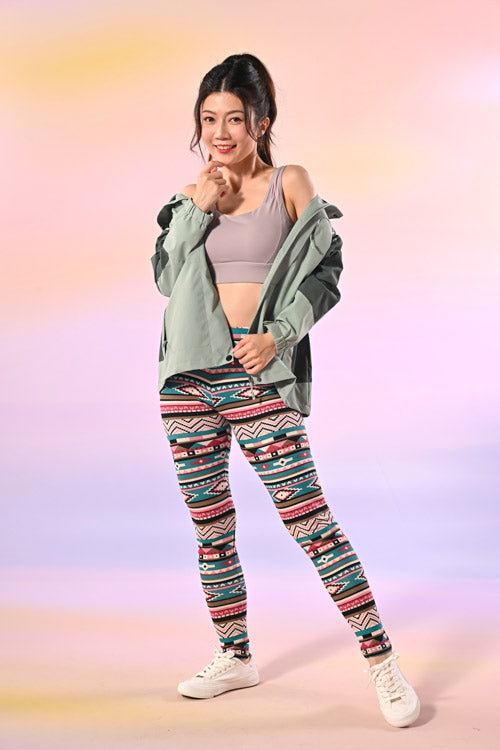 一位女士身穿淺灰色運動胸罩、花紋緊身褲、白色運動鞋，肩上掛著一件綠色外套，站在粉色漸變背景下微笑。.