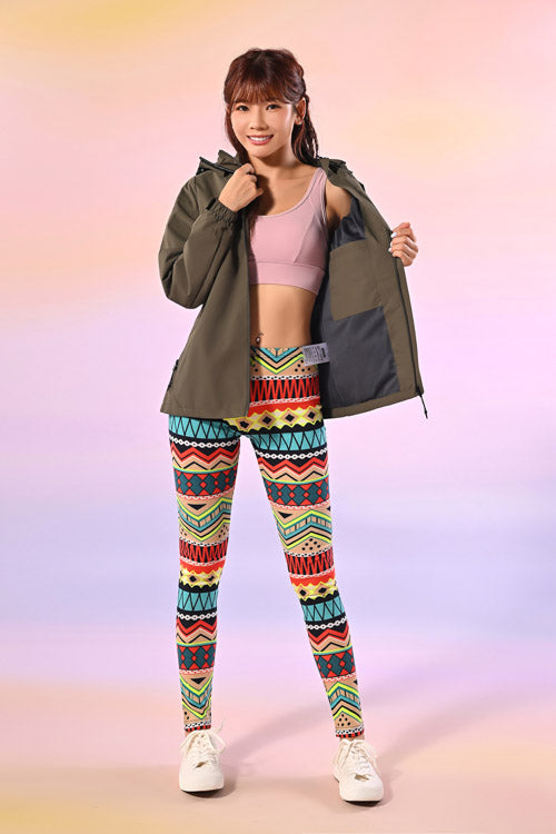 一位面帶微笑的女士站在粉色背景的工作室中，身穿粉色運動胸罩、開放式橄欖綠外套、彩色幾何圖案緊身褲和白色運動鞋。.